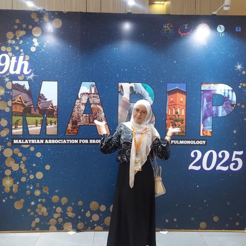 Ariantika in conference lanyard gesturing toward the 9th MABIP 2025.