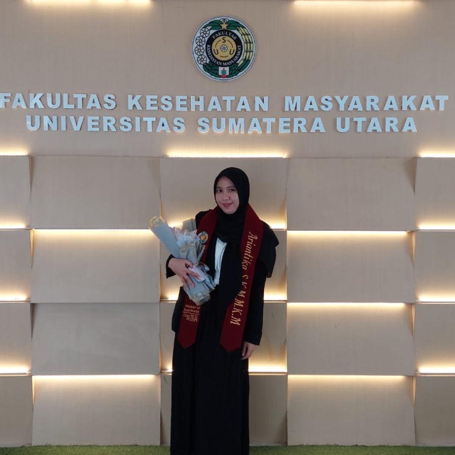 Ariantika in graduation gown and master's sash holding a bouquet in front of the Faculty of Public Health wall at Universitas Sumatera Utara