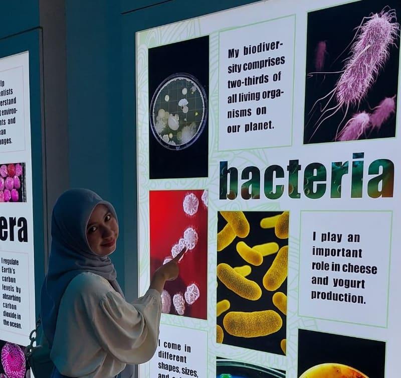 Ariantika smiling while pointing at a museum exhibit panel about bacteria.